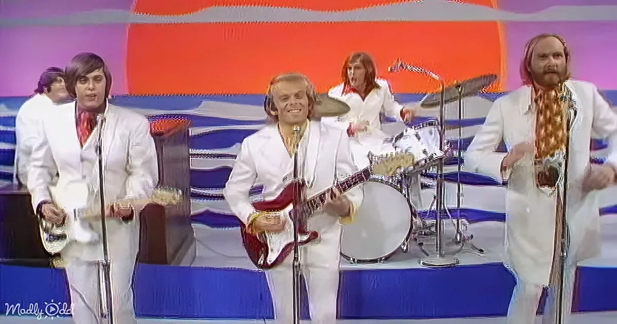 A smiling man in a white suit plays a red guitar on stage with a colorful background.