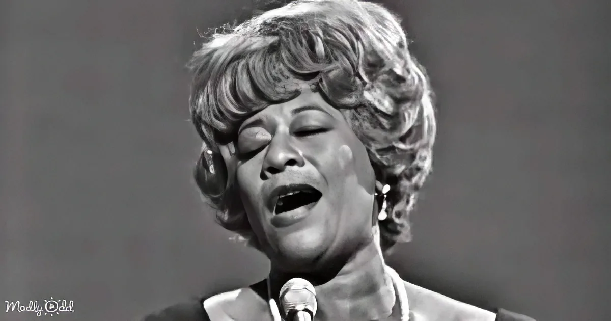 A woman with short, curly hair sings passionately into a microphone, her eyes closed, in a black-and-white image.