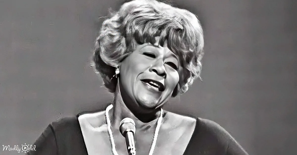 A woman with short, styled hair is singing into a microphone, wearing a formal outfit.
