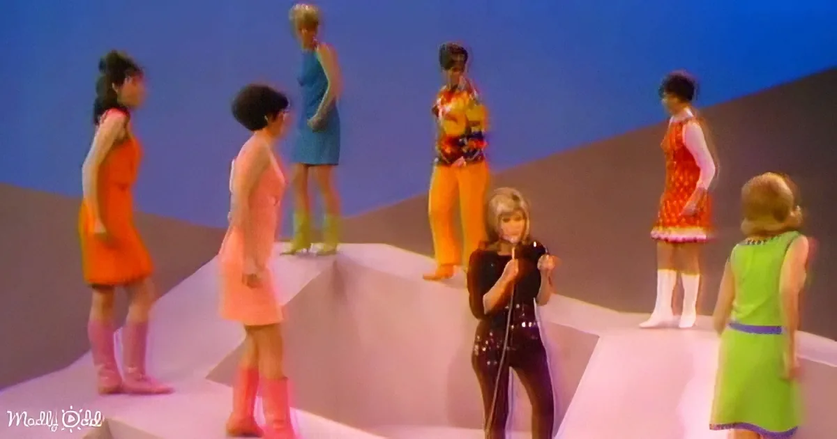A performer in a black outfit sings on stage, surrounded by dancers in colorful outfits.