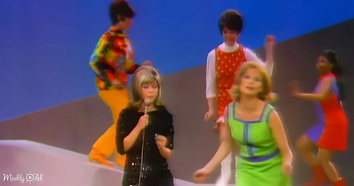 A woman in a black outfit sings into a microphone on stage, surrounded by dancers in colorful outfits.