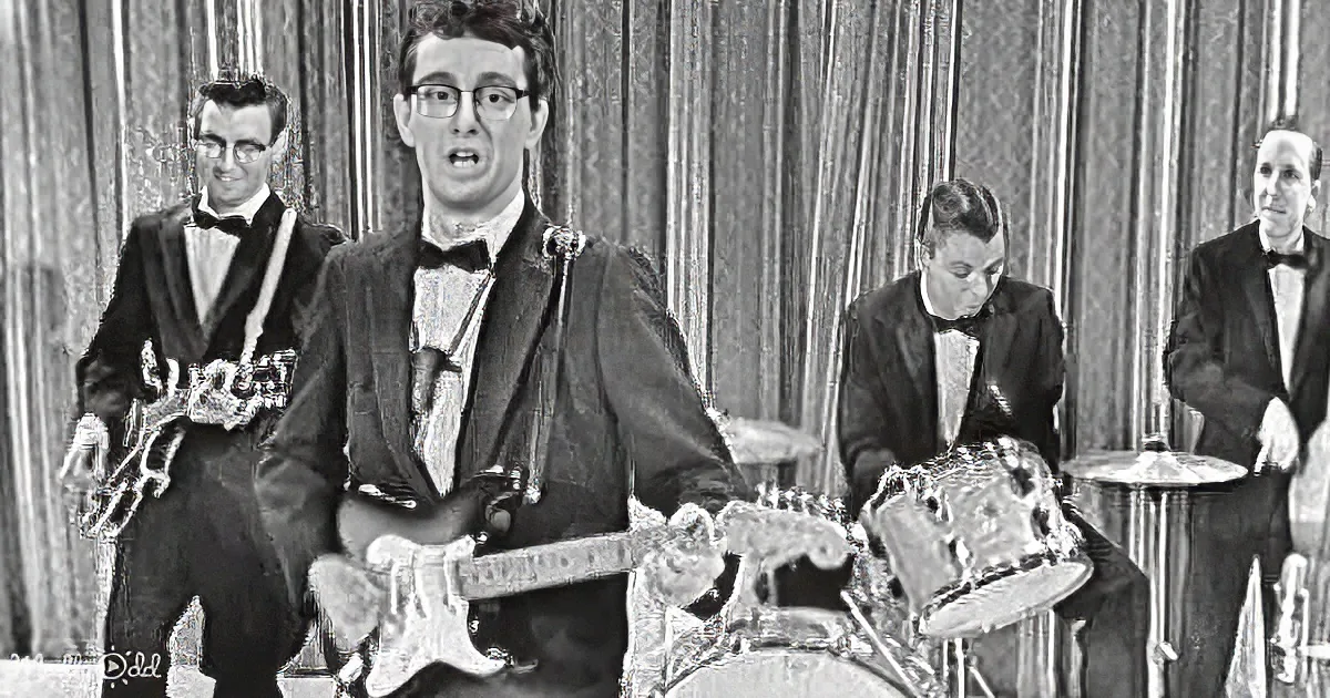 A man in glasses plays guitar, surrounded by a band in formal attire, with a curtain backdrop.