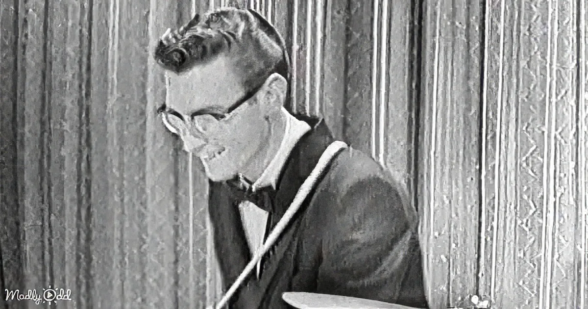 A man in a suit and bow tie sits at a table, holding a stick, with a wooden-paneled wall behind him.