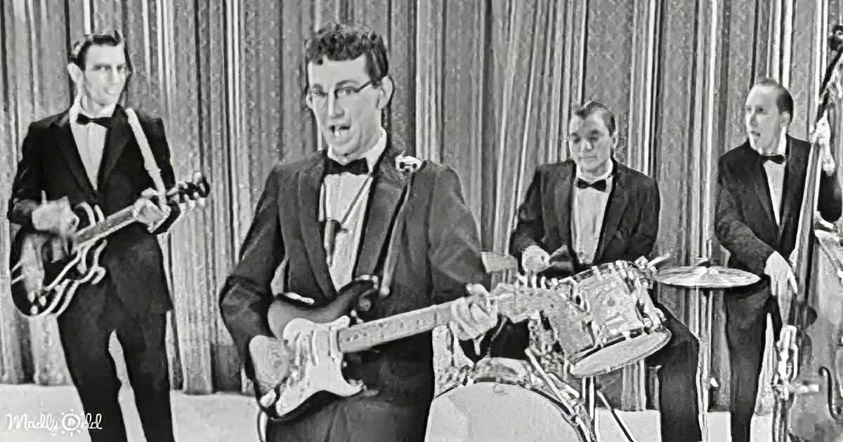 A man in a tuxedo plays a guitar while a band in formal attire performs.