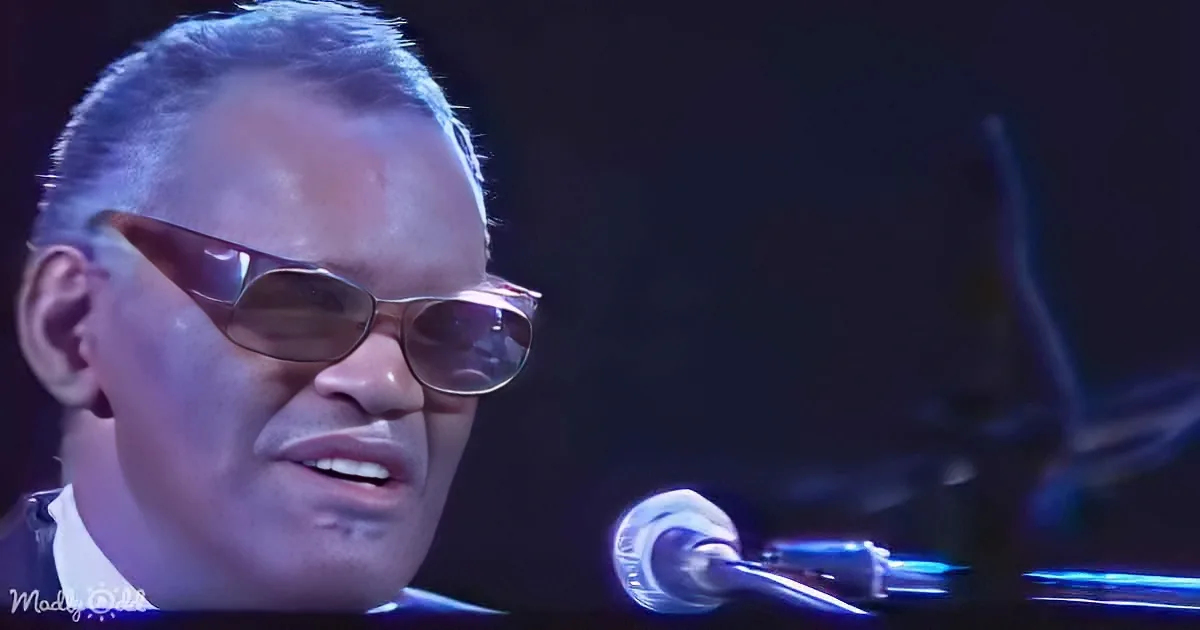 A man wearing sunglasses and a suit sings into a microphone, with a dark background and a hint of blue light.