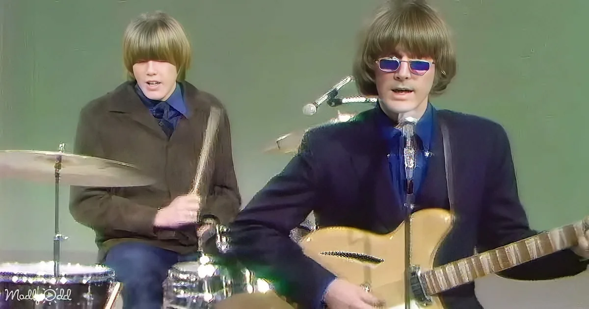 A man in a suit plays guitar while another in a brown jacket plays drums.