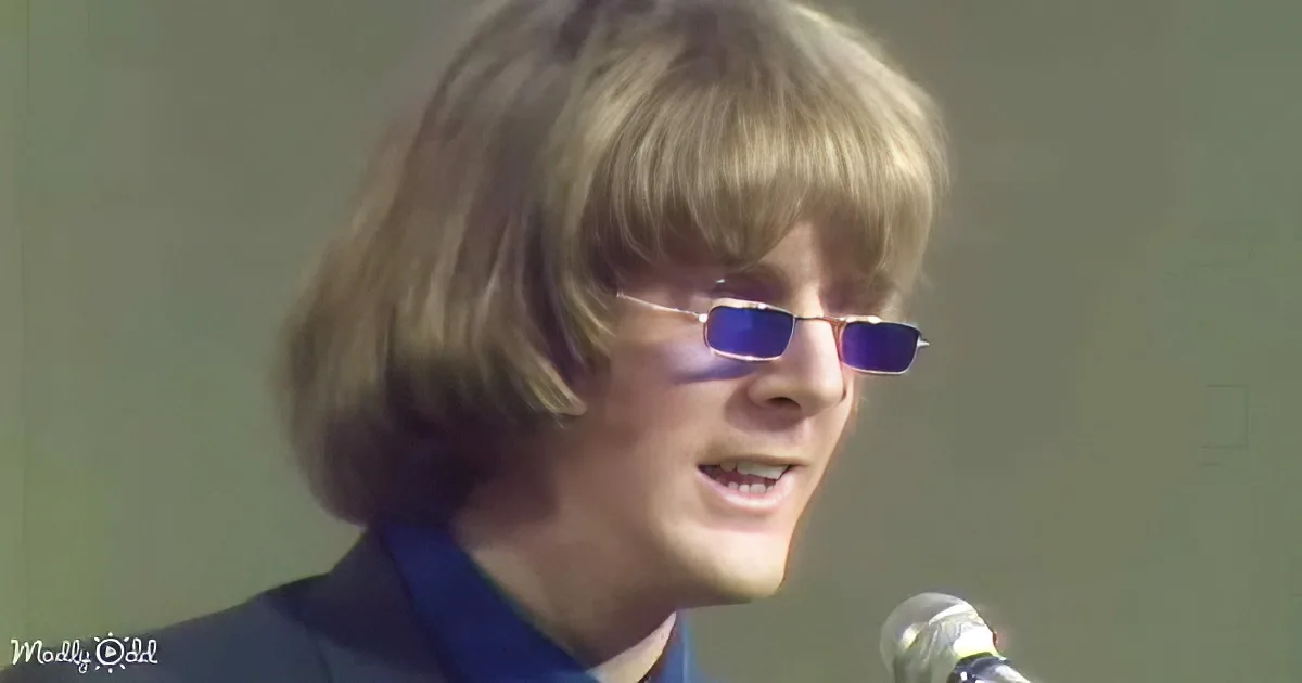 A person with shoulder-length hair and sunglasses, wearing a dark suit, stands in front of a microphone.