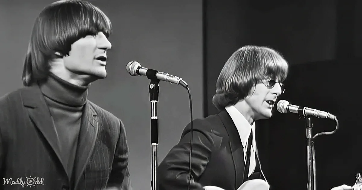 Two individuals with long hair, wearing suits, singing into microphones, one wearing glasses.