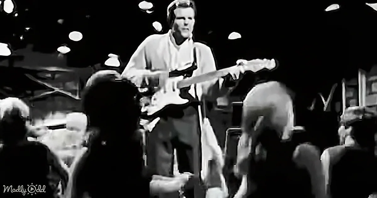 A man plays guitar on stage, surrounded by a crowd, in a black-and-white image.