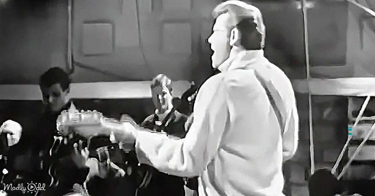 A man in a white shirt plays a guitar on stage, with a band in the background.