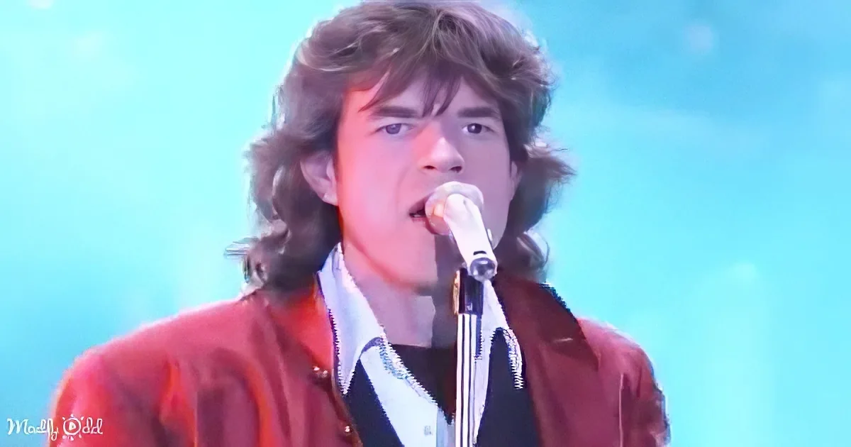 A person with long hair, wearing a red jacket, sings into a microphone against a blue background.