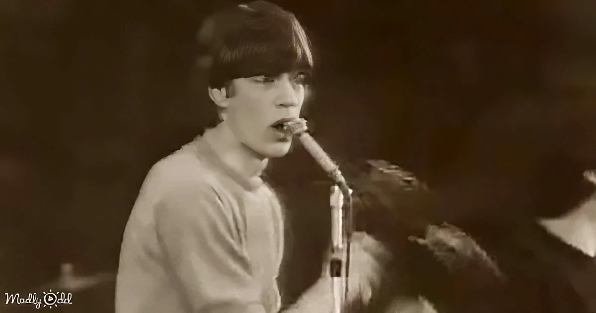 A person in a white shirt sings into a microphone, with a dark background and another figure partially visible.