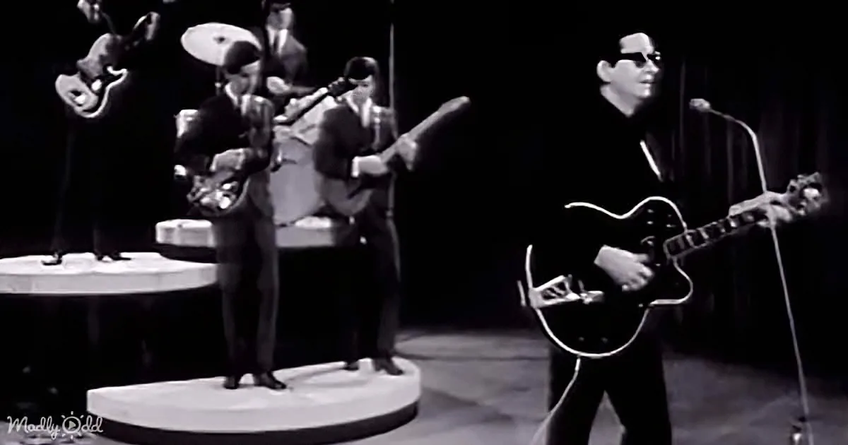 A guitarist in a dark suit plays a black electric guitar on stage with other musicians.