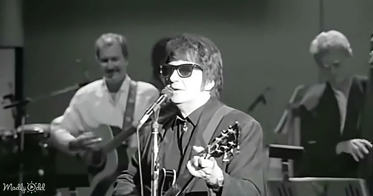 A man in sunglasses plays guitar, singing into a microphone, with two other musicians in the background.