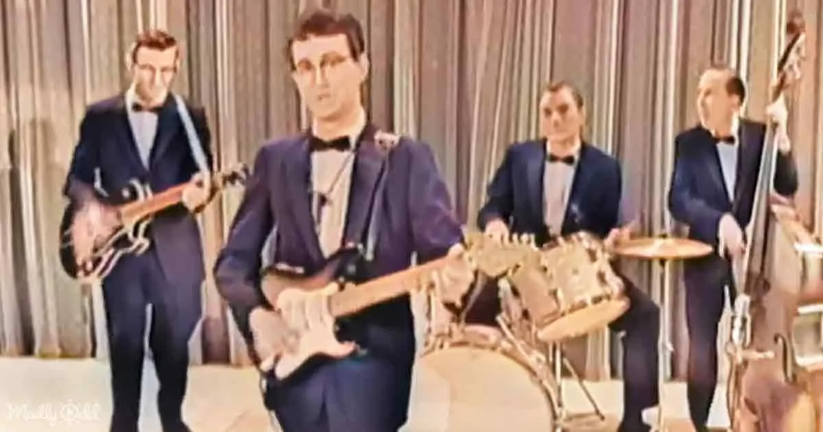 A man in a blue suit plays a Stratocaster guitar on stage with a band.