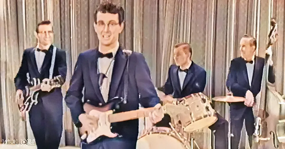 A man in a blue suit plays guitar, surrounded by other musicians in formal attire.