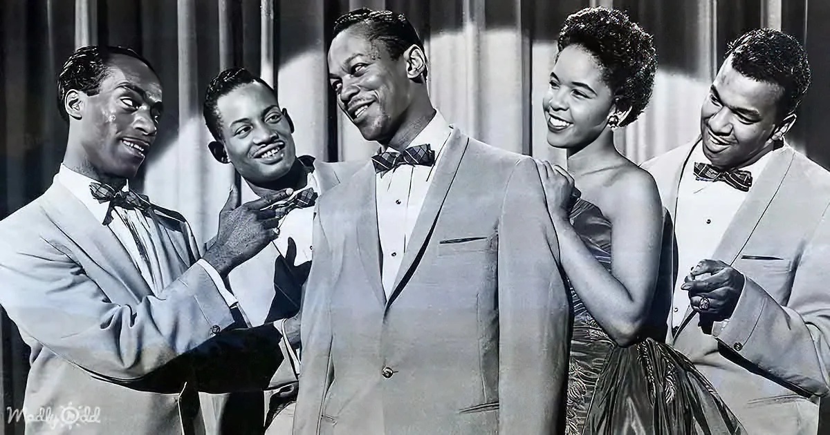 A man in a suit is being adjusted by another man, with a woman and two other men smiling around them.
