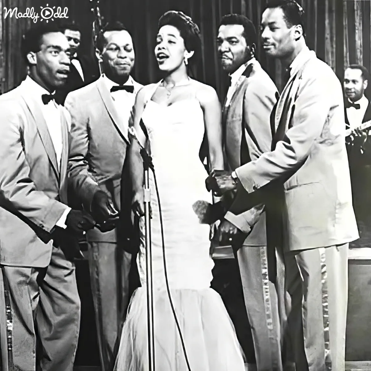 A woman in a white dress sings on stage with a group of men in suits.