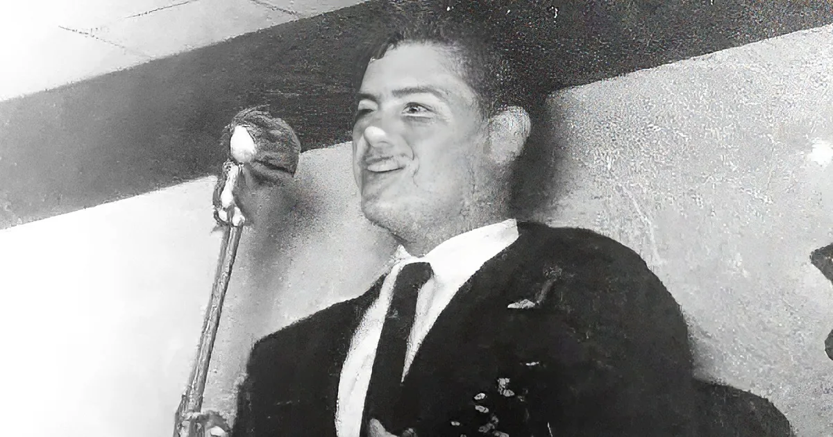 A man in a suit and tie stands beside a microphone, smiling, in a black-and-white photo.