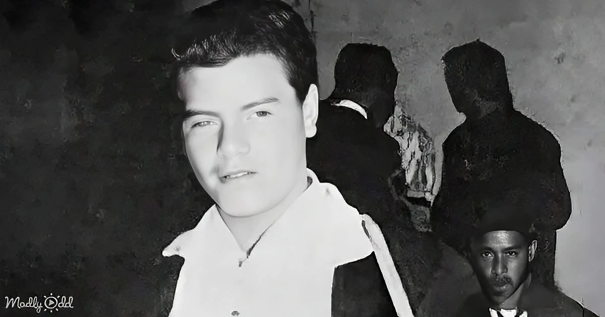 A man in a white shirt stands in front of a dark background, with silhouettes of other people behind him.