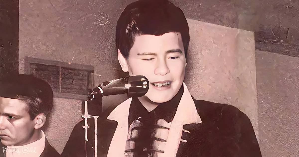 A person in a black and white outfit sings into a microphone, with another person partially visible to the left.