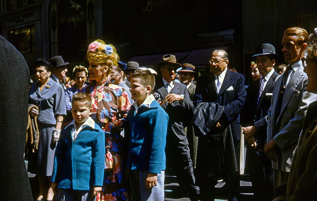 A boy in a blue coat stands among a crowd of people, some in suits and hats, on a city street.
