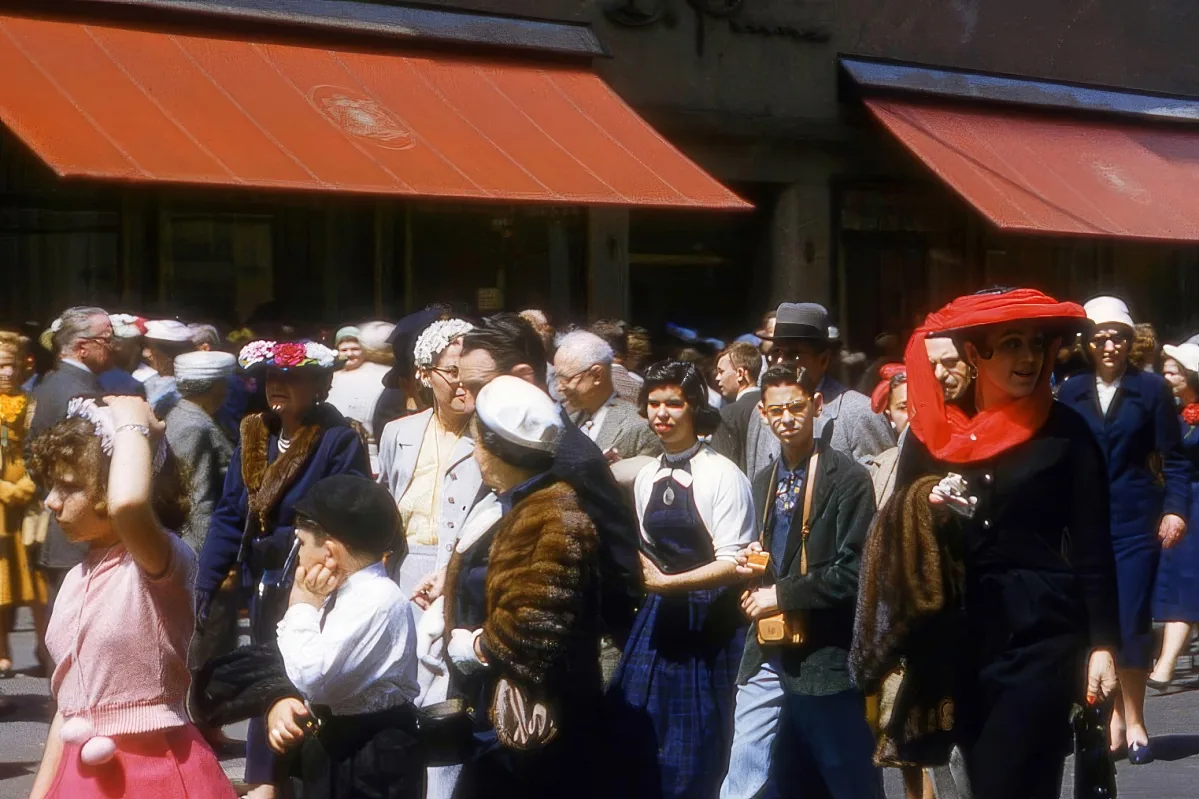 A bustling crowd of people in vintage attire, with red awnings and shops in the background.