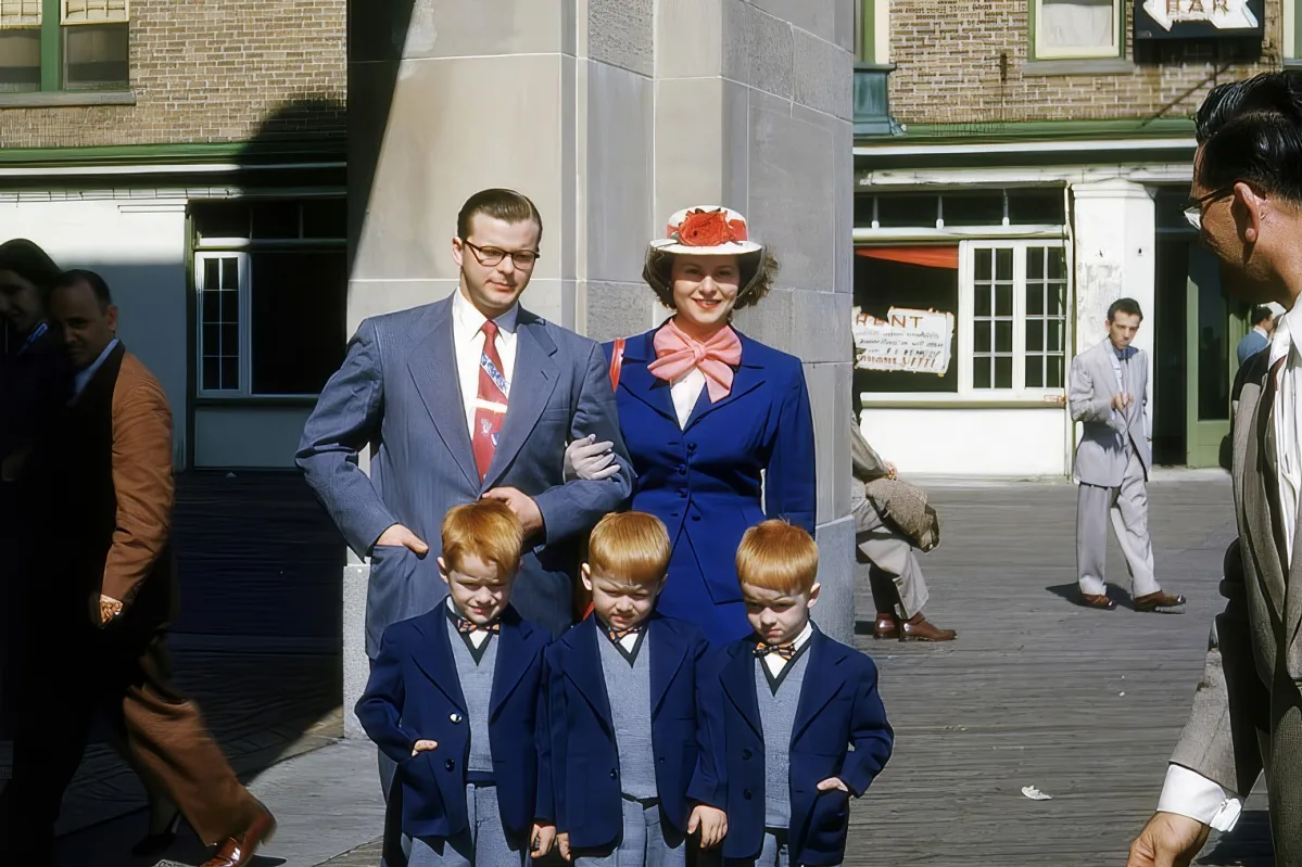 A family in matching blue suits and hats stands together on a sunny day, with a man in a brown suit and a woman in a pink...