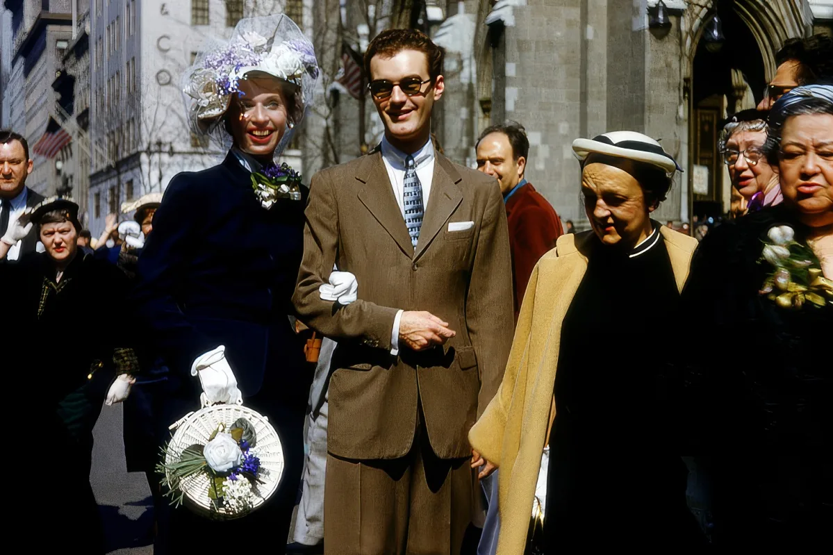 A man in a brown suit and sunglasses stands with a woman in a floral hat, smiling amidst a crowd of people in a city setting.