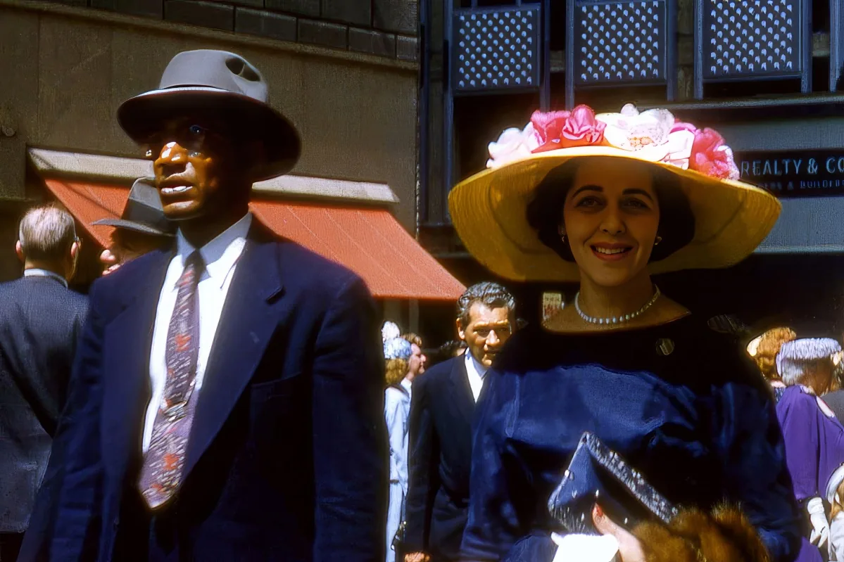 A man in a suit and hat stands beside a woman in a blue dress and floral hat, both in a bustling street scene.