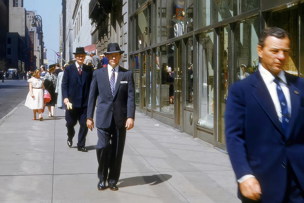 A man in a suit and hat walks down a city sidewalk, surrounded by buildings and pedestrians.