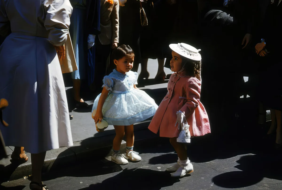Two young girls in dresses and hats stand on a street, surrounded by adults, under bright sunlight.