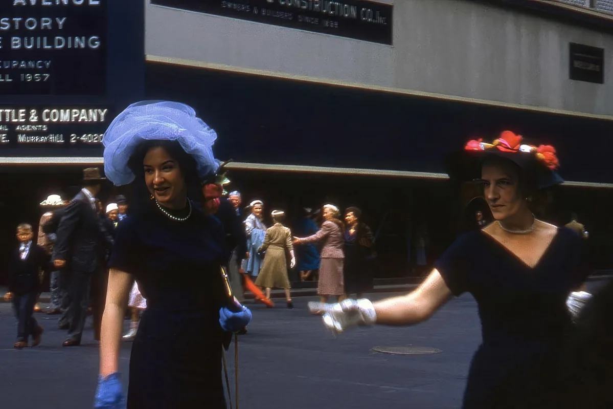 A woman in a black dress and blue hat is dancing on a city street, surrounded by other people.