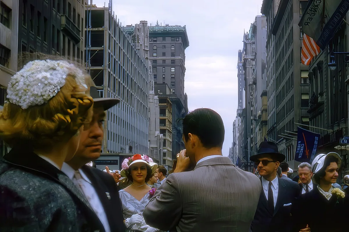 A woman in a white dress and floral hat is surrounded by people in a bustling city street with tall buildings.