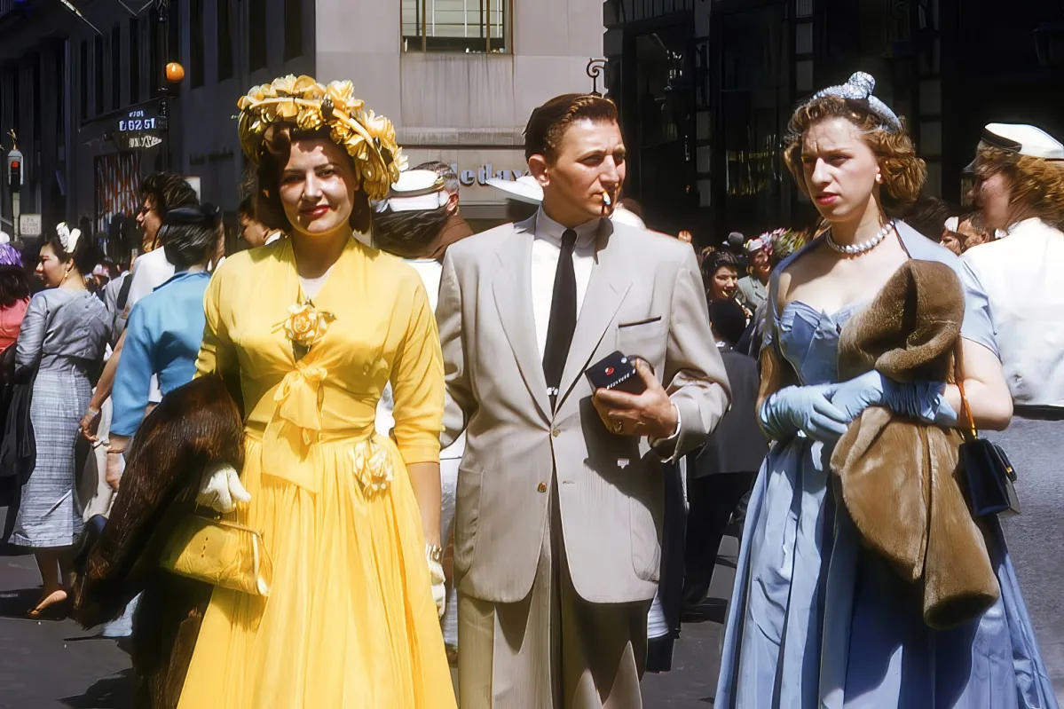 A woman in a yellow dress with a floral headpiece walks alongside a man in a light suit and a woman in a blue dress with...