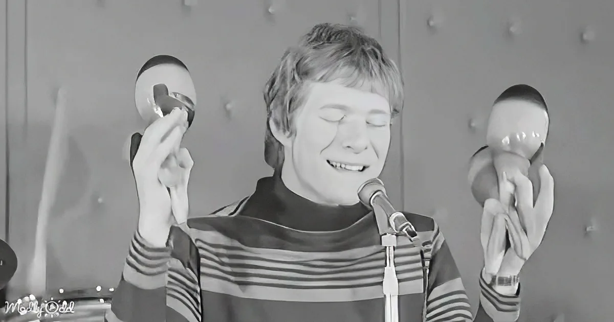 A person in a striped shirt holds a maraca and a doll, singing into a microphone.