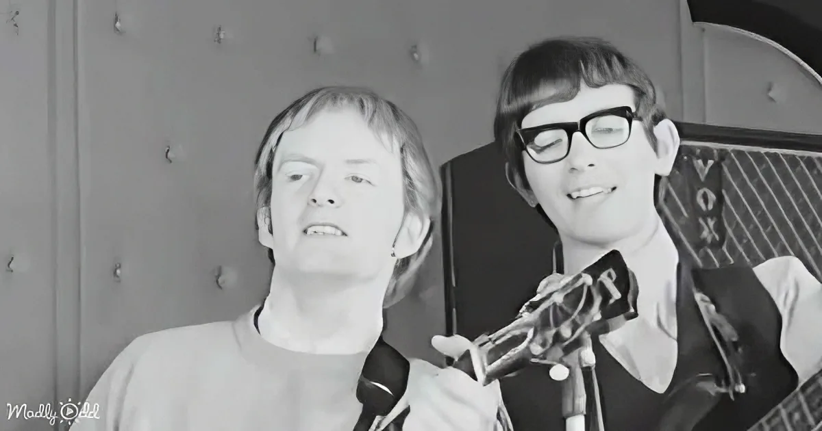 Two individuals, one playing guitar, both singing into microphones, in a black-and-white setting.