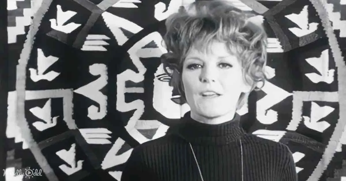 A woman with short, curly hair in a black turtleneck stands in front of a black-and-white abstract background.