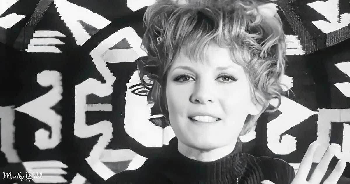 A woman with short, curly hair and a black top stands in front of a black and white abstract background.