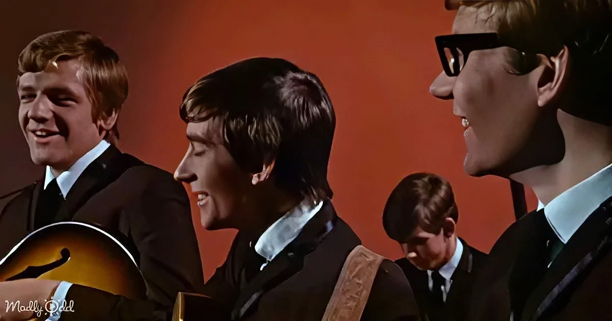 Four young men in suits, one playing guitar, smiling, against a red background.