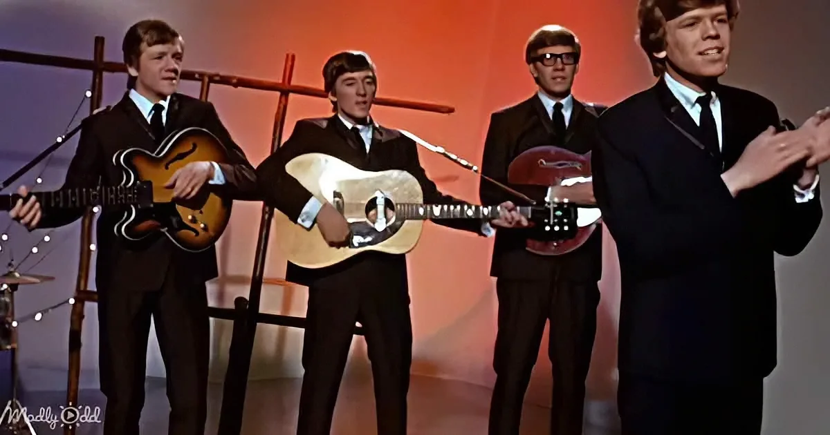 Four young men in suits play guitars on stage, one clapping, under a warm, orange-lit backdrop.