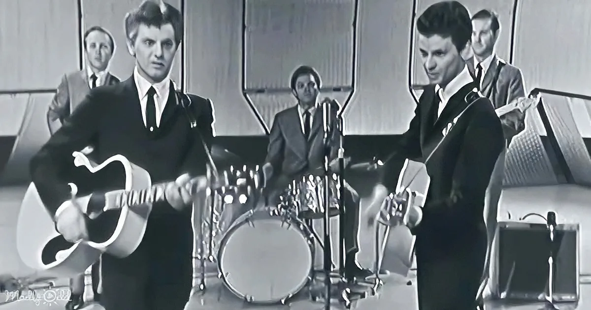 A man in a suit plays guitar on stage with a band, including a drummer and a guitarist.
