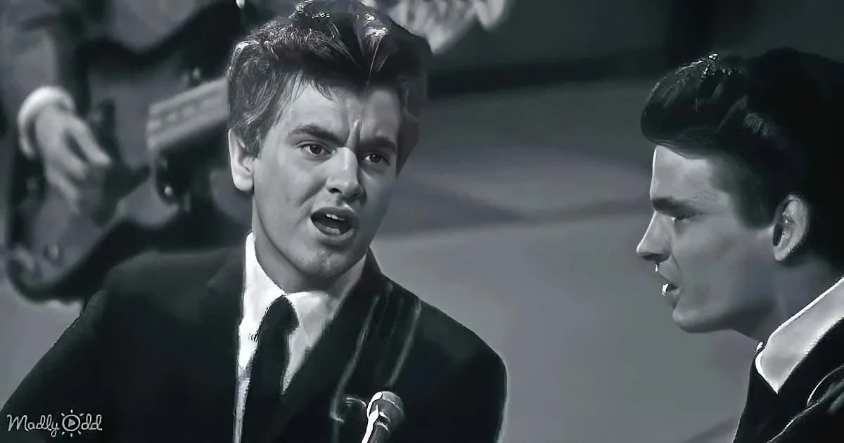 A man in a suit sings passionately into a microphone, with another person nearby, both in a performance setting.