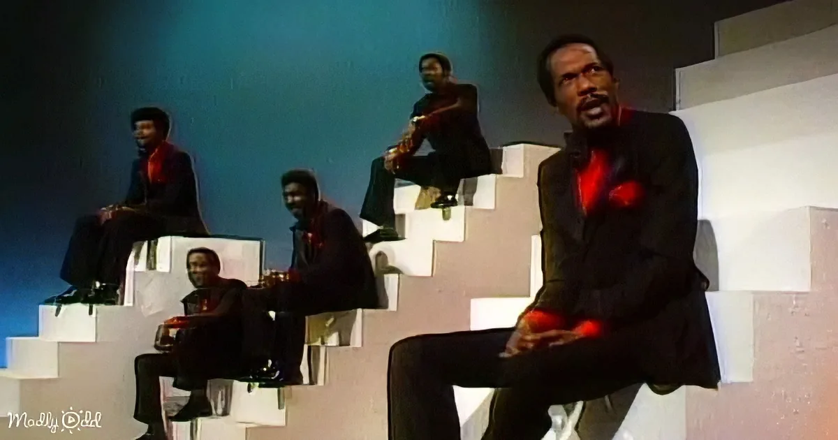 A man in a black suit sits on a white block, surrounded by others in similar attire on a stage with a blue and white...