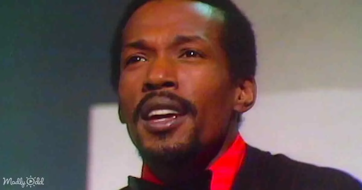 A man with a mustache and beard, wearing a black suit with a red shirt, appears to be speaking or singing.