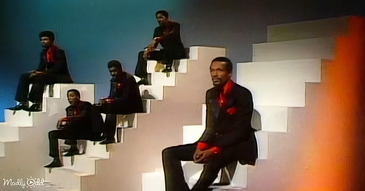 A man in a suit sits on a white block, surrounded by others in similar attire on a staircase-like structure.