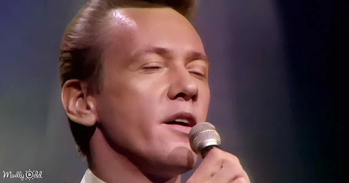 A man with slicked-back hair sings into a microphone, eyes closed, in a dimly lit setting.