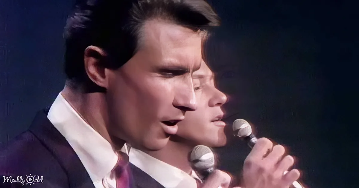 A man in a suit sings into a microphone, with another person partially visible behind him.