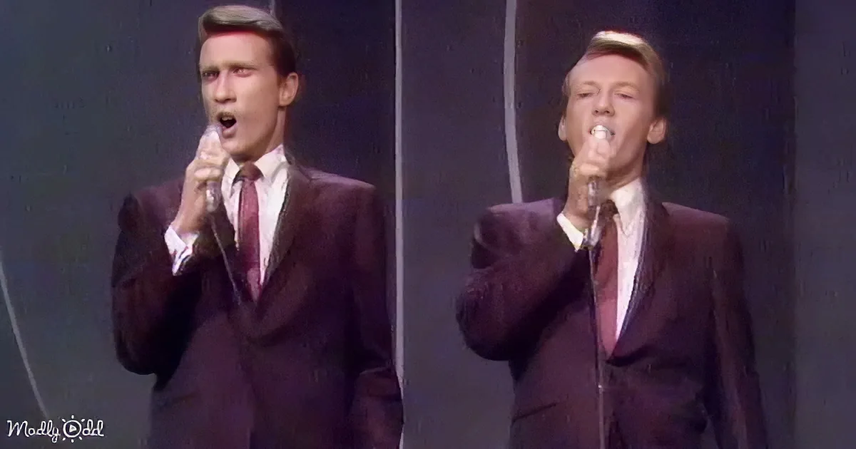 Two men in suits sing into microphones on stage, one with a red tie.