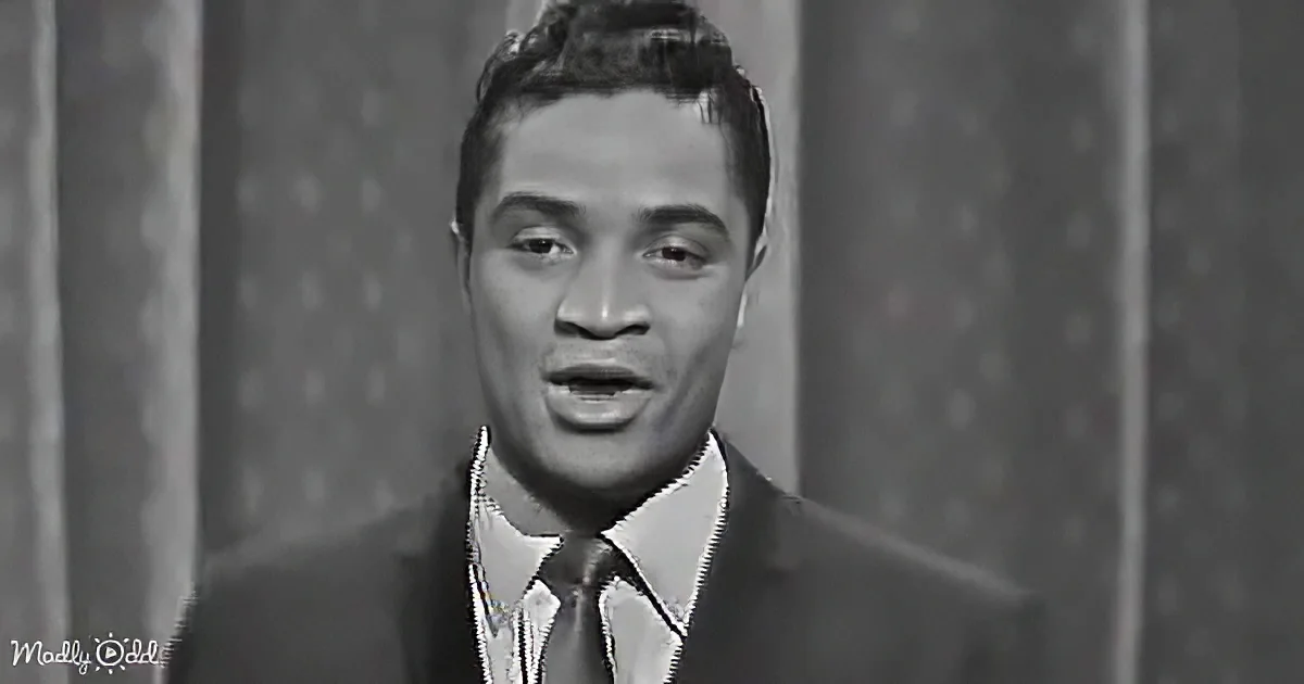 A man in a suit and tie, possibly singing or speaking, with a curtain backdrop.